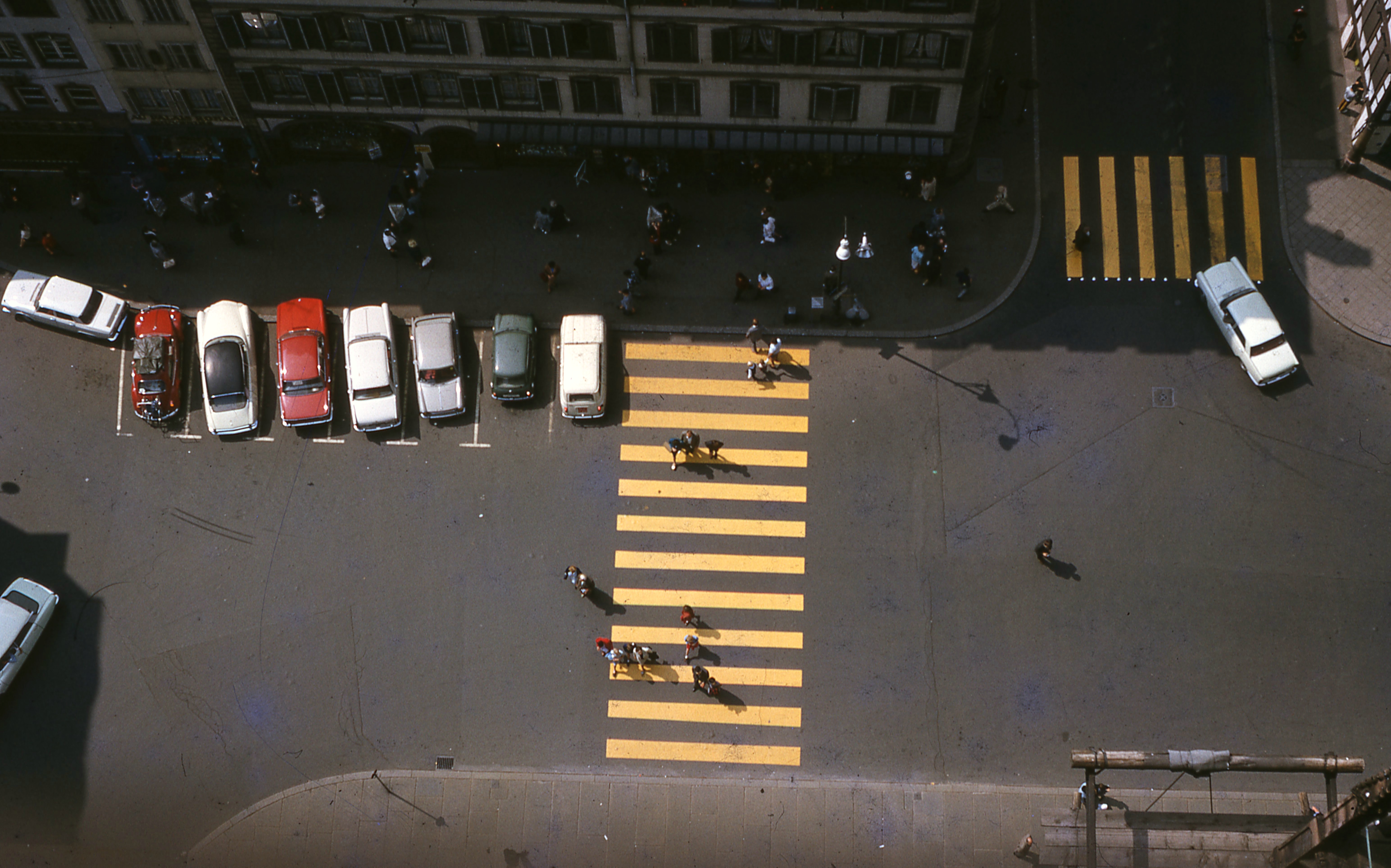 Aerial photo of a yellow zebra crossing and pedestrians. A few cars are parked in front of the building opposite.