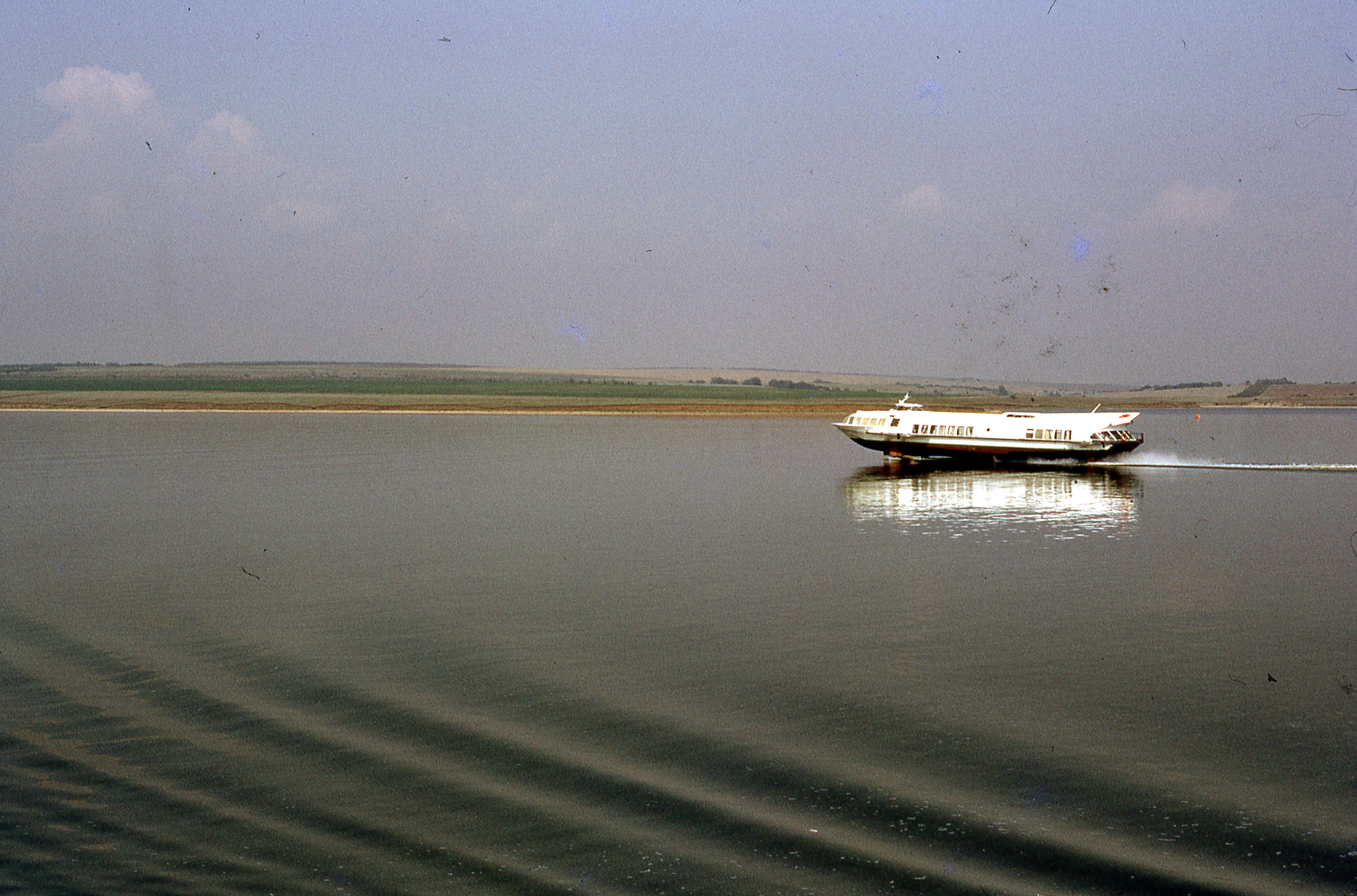 A flat motor boat is travelling down the river, and reflected in the water. The landscape behind is flat.