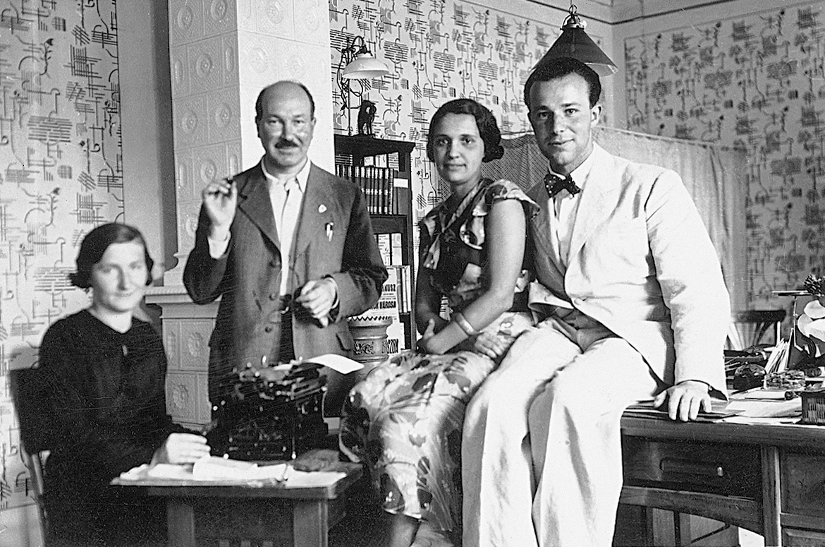 Portrait of four people in an office, one woman sits at a desk in front of a typewriter, one man is standing, and a man and a woman are sitting on a table. The room has abstract wallpaper and a large tile oven.