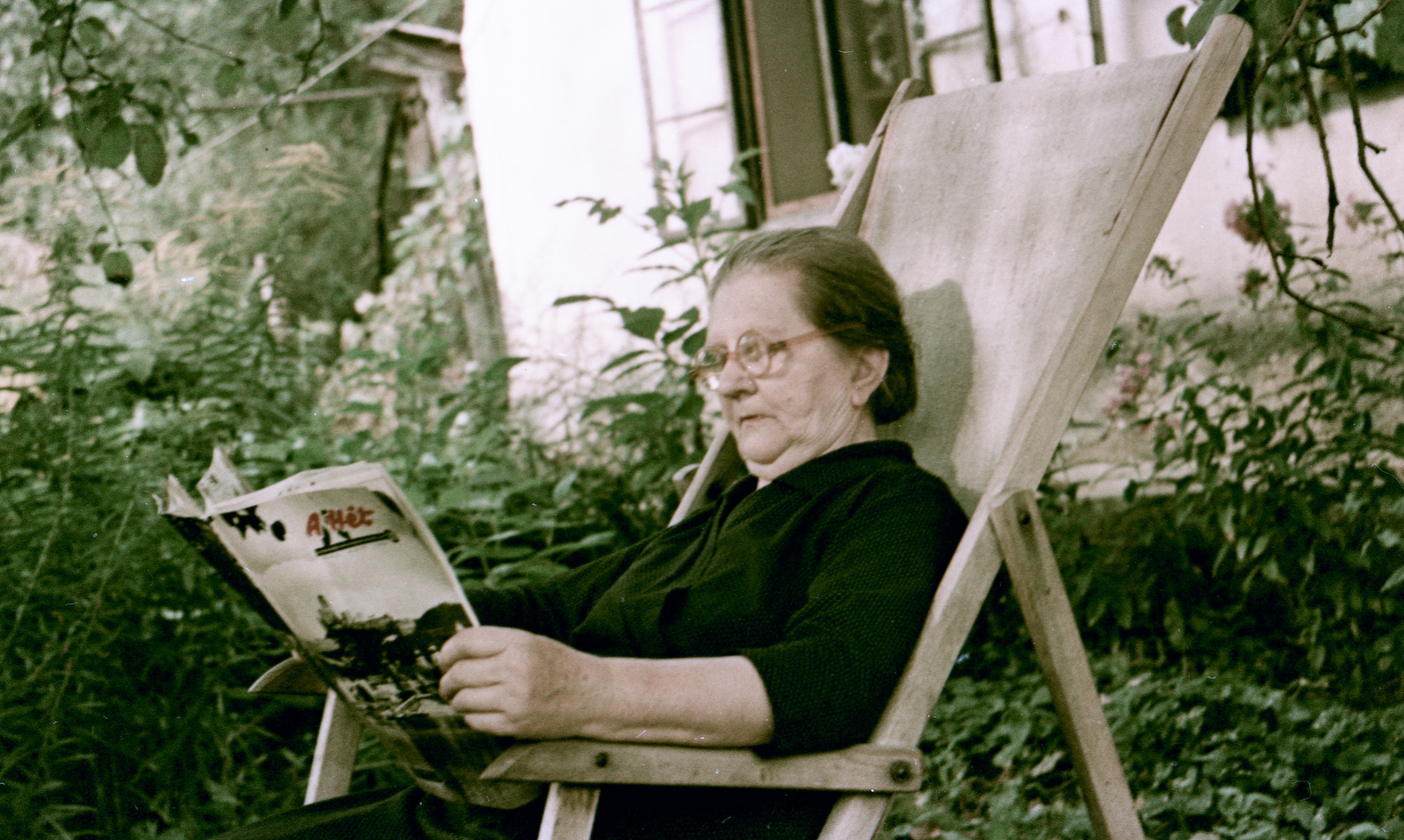 In a garden, an elderly woman sits in a canvas chair reading a magazine.