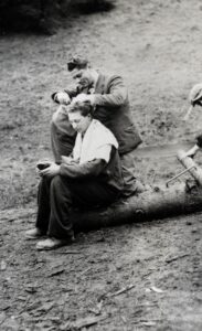 A man in a suit jacket is carefully cutting the hair of another man sitting on a one end of a felled tree trunk with a towel around his shoulders. Behind them is an incline with pine trees. Three more forced labourers are partially visible at the edges of the photo.
